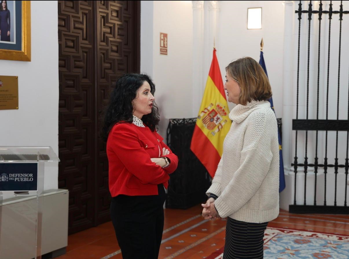 La presidenta de la AINODI y la adjunta segunda del Defensor del Pueblo están de pie conversando entre ellas. Tienen detrás, en el centro, las bandera de España y de la UE, a la derecha una puerta de rejería y a la izquierda otra de madera labrada de gran altura