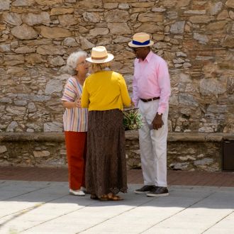 Dos mujeres y un hombre, todos de edad avanzada, conversan en círculo en un día soleado y con ropa de entretiempo. El hombre y una de las mujeres llevan sombreros de paja