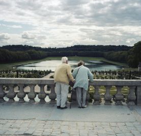 Pareja de personas mayores, mujer y hombre, que están de espaldas mirando desde una balaustrada a un jardín con un lago en el centro