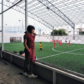 Una niña en primer plano vestida con ropa roja de calle contempla desde la grada un partido de futbol infantil masculino, equipado también de rojo,