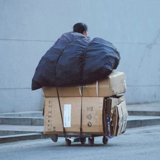 Una persona tira de un carro cargado con cajas de cartones en su parte inferior, y en la superior, de un gran bulto envuelto en una lona azul