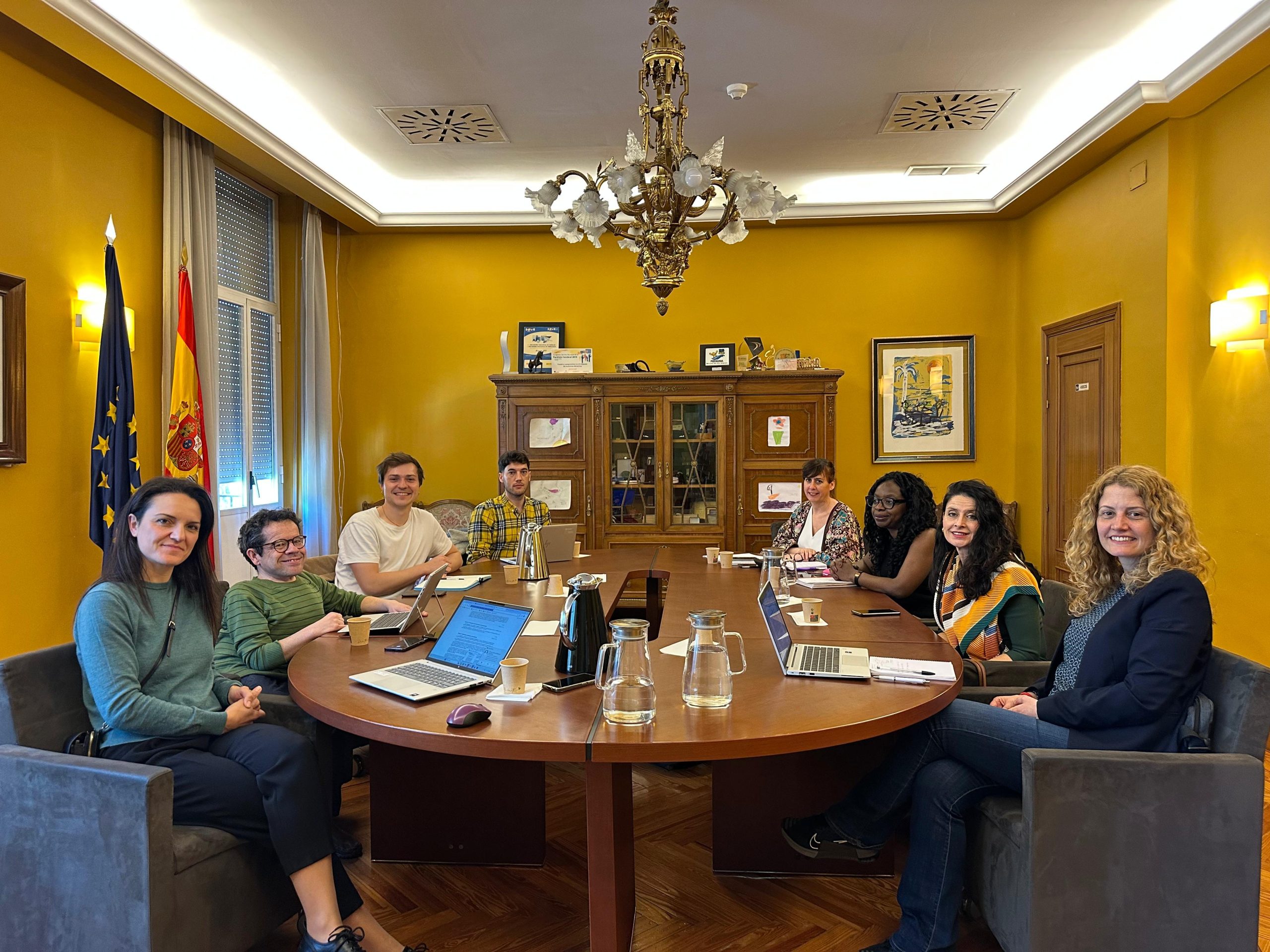 La presidenta de la AINODI y el director del Real Patronato sobre Discapacidad en una mesa de reuniones junto a seis personas de sus respectivos equipos. A la izquierda, las banderas de España y de la UE