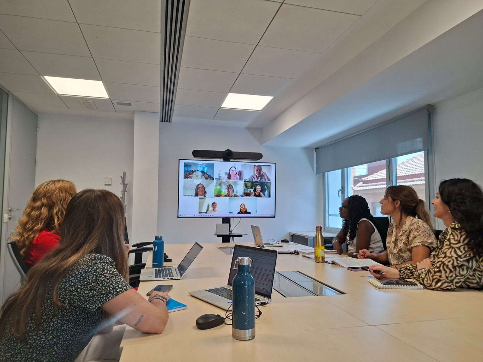 La presidenta de la Autoridad independiente y el equipo técnico de la AINODI en una mesa de reuniones de espaldas mirando a una pantalla en la que hay personas de Espai Unitari en una videoconferencia